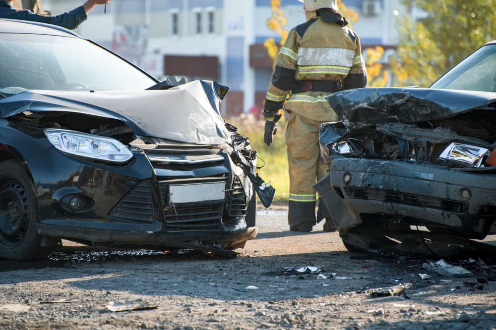 Two heavily damaged cars after a collision, with a firefighter walking in the background.