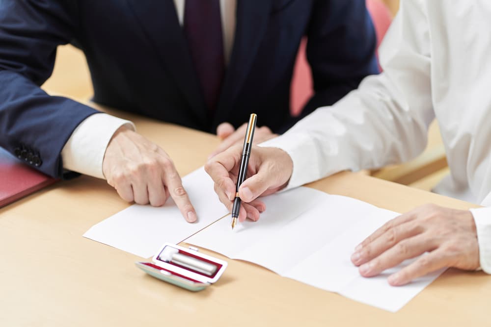 Two people at a table, one directing where to sign a document, close-up view.