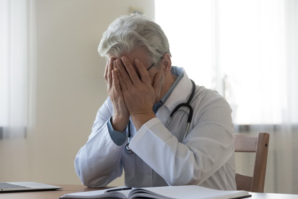 Distressed elderly doctor sitting at desk, covering face with hands in a somber moment.