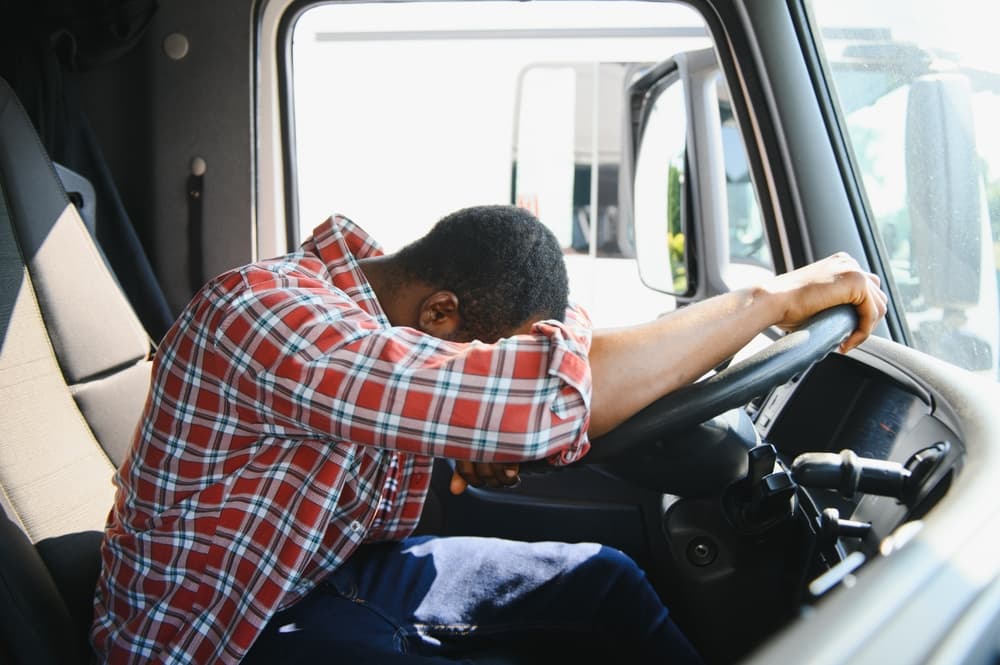 Distressed truck driver resting head on steering wheel, appearing exhausted or upset.