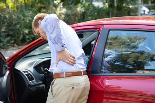 A man standing outside of a red car, holding his lower back in pain, implying a back injury potentially caused by a car accident.