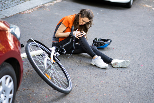 A woman in an orange shirt and black leggings sits on the ground next to a fallen bicycle, holding her injured knee, with a car in the background.