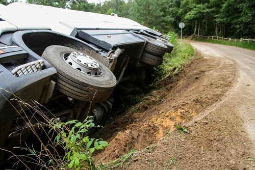 A large truck overturned on the side of a rural road, with its wheels in the air and the cabin partially off the road, indicating a severe accident.