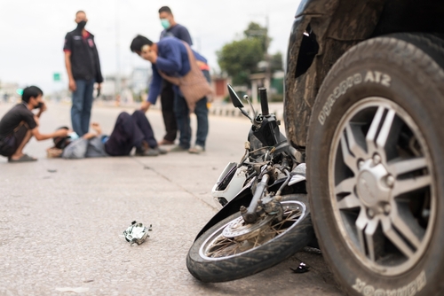 A damaged motorcycle on the ground near the tire of a car after an accident, with people gathered in the background, some attending to an injured person.