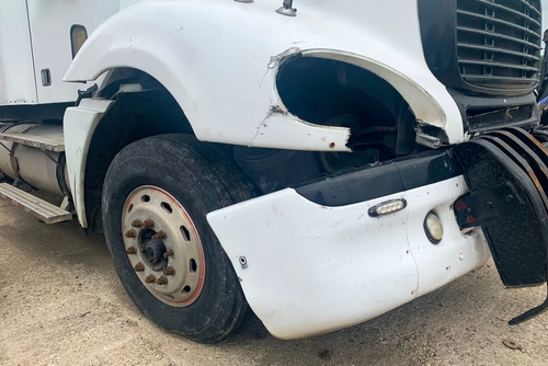 A close-up view of a damaged white truck with significant front-end damage, including a broken headlight and dented fender, likely caused by an accident.