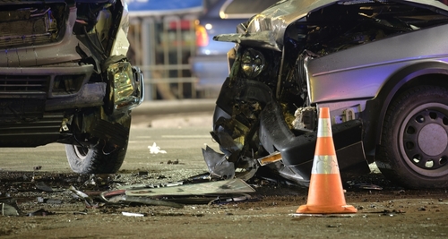 A close-up view of a serious car accident scene showing two heavily damaged vehicles at night with a traffic cone in the foreground.