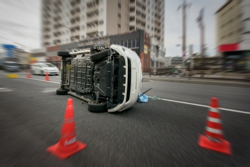 A white vehicle on its side following a rollover accident on a city street, surrounded by orange traffic cones with blurred buildings in the background.