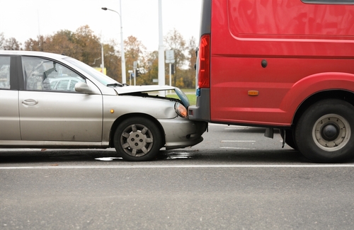 A silver car involved in a rear-end collision with a red van on a road. The front of the silver car is visibly damaged where it hit the back of the van.