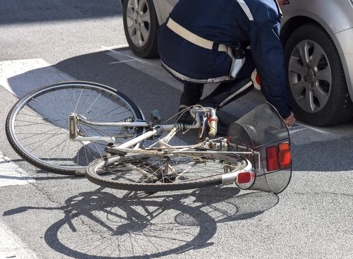 A cyclist in a blue jacket inspects a fallen bicycle next to a parked car after a collision, with the bike lying on the road.