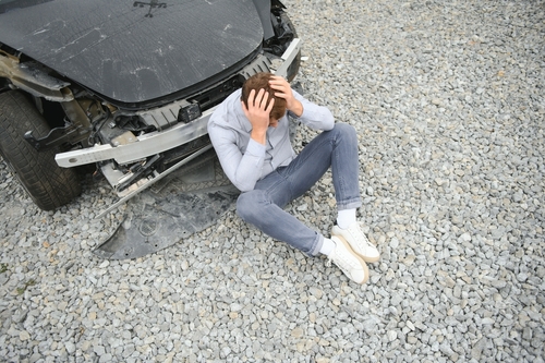 Man sitting on the ground, holding his head in distress in front of a heavily damaged car, indicating the aftermath of a car accident.