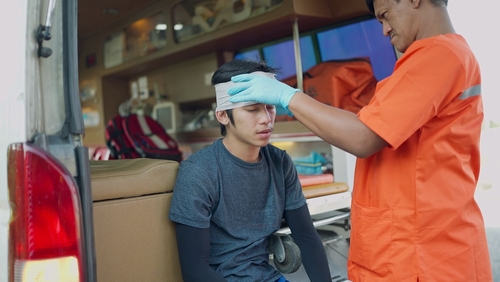 A paramedic attending to a young man with a head injury, wrapping a bandage around his forehead while sitting in an ambulance.