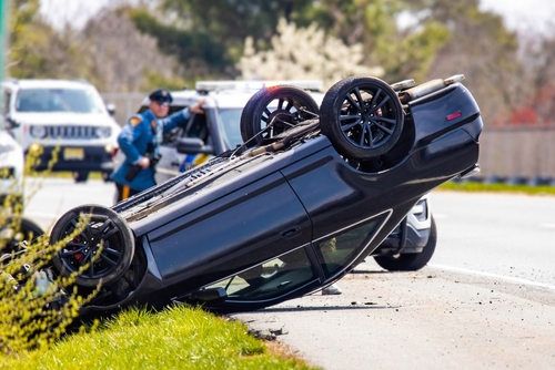 A black vehicle overturned on its roof after a rollover accident on a roadside, with police officers and patrol cars in the background.