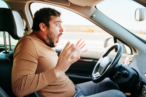 A man driving a car with a shocked expression on his face, raising his hands in alarm as if reacting to an unexpected situation on the road.