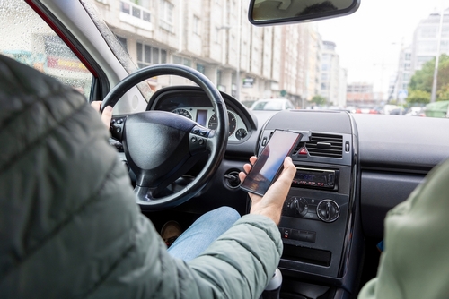 A driver holding a smartphone in one hand while steering the car with the other, suggesting distracted driving in an urban setting.