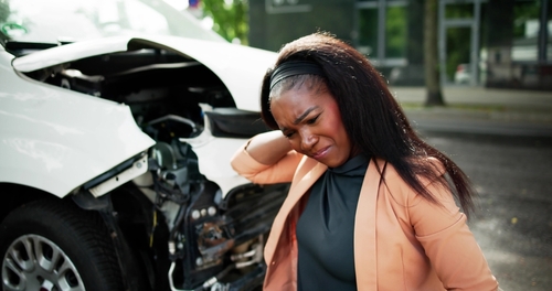 Woman holding her neck in pain while standing near a car with significant front-end damage after an accident