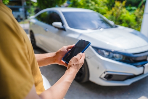  A person holding a smartphone in front of a parked white car, possibly capturing or reviewing a photo of the vehicle.