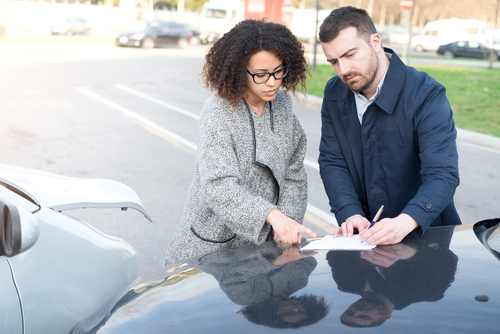 A man and a woman stand beside two vehicles that appear to have been in a minor car accident. They are reviewing and signing a document on the hood of one of the cars, likely an insurance or accident report form.