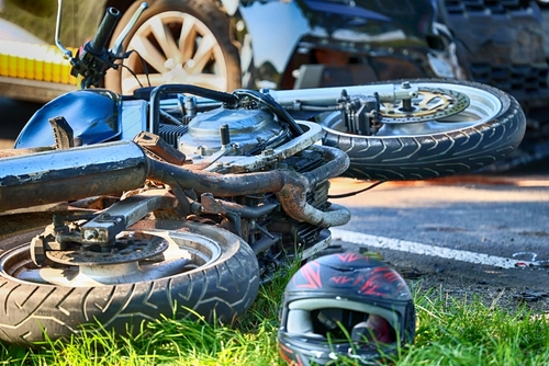 Close-up of a damaged motorcycle lying on the road after an accident, with a helmet resting nearby on the grass.