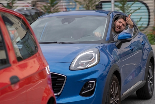 Angry driver in a blue car leaning out of the window, making an aggressive gesture toward another vehicle, illustrating road rage.