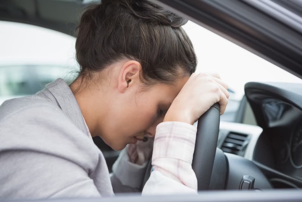 Woman asleep at the wheel of her parked car, head resting against the window, with dim interior lighting and a quiet street scene outside.






