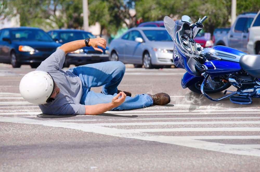 A motorcycle rider lies injured on the road after a crash, while their bike skids across the pavement toward a busy intersection.