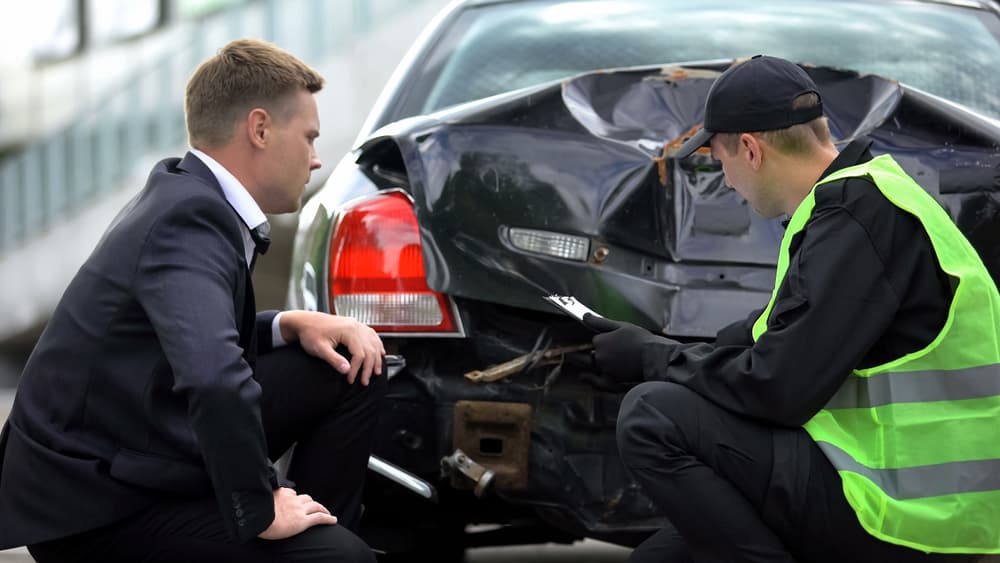 Police inspector examining the damage and filling out a car accident report while a stressed driver looks on.