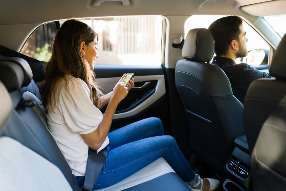 A side view of a woman seated in the back seat of a car, holding her smartphone and interacting with a rideshare app, with soft natural light illuminating her face.