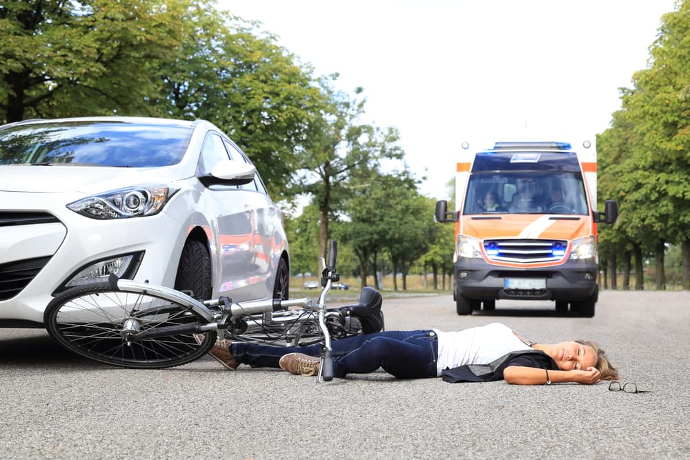 A young woman lies injured after a bicycle accident, with an ambulance arriving at the scene.






