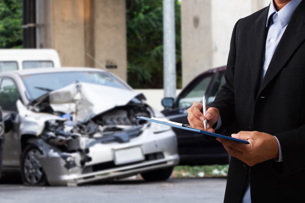 An insurance officer writes on a clipboard while an insurance agent carefully examines a silver car and a black car involved in an accident. 