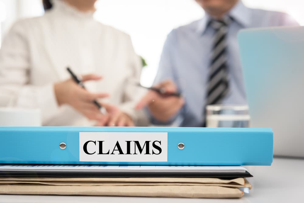 A claims document folder is on the table as a lawyer discusses the insurance claim policy with a board manager in a meeting room.