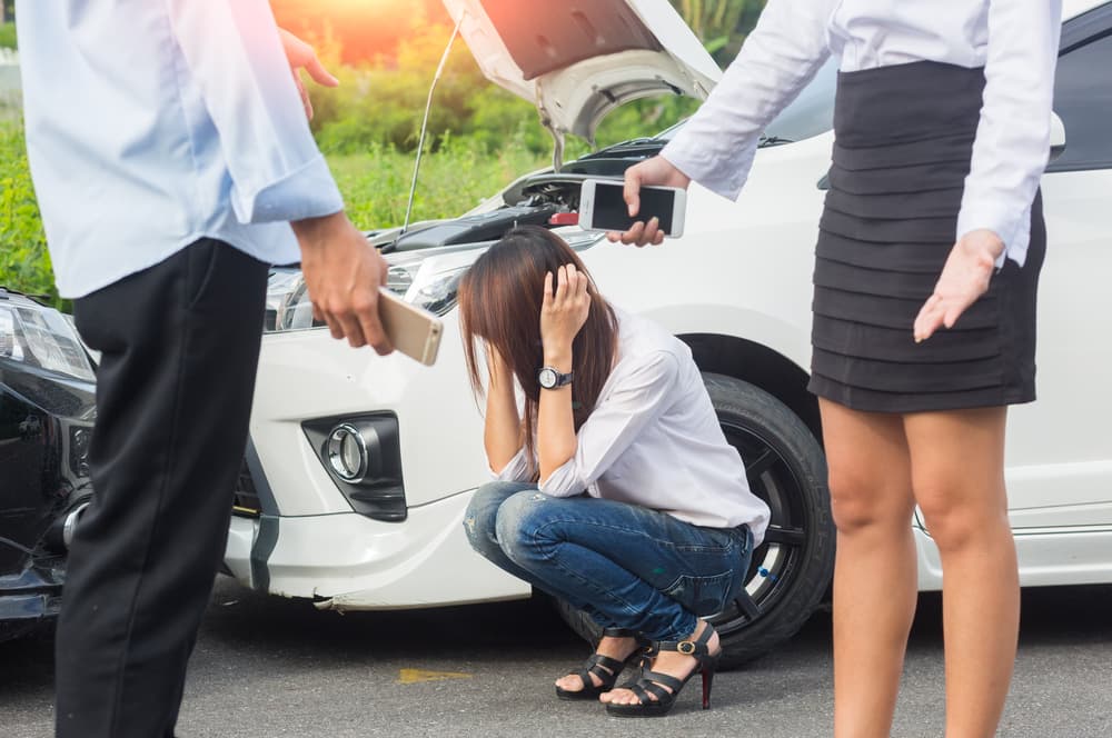 An Asian woman, the driver, stands in front of a damaged car after a collision on a city road, appearing concerned as she assesses the accident scene.






