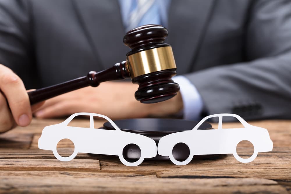 A judge's hand is seen striking a gavel near paper cars placed on a wooden table, symbolizing legal decisions related to automobile cases such as accidents, insurance disputes, or vehicle ownership issues.