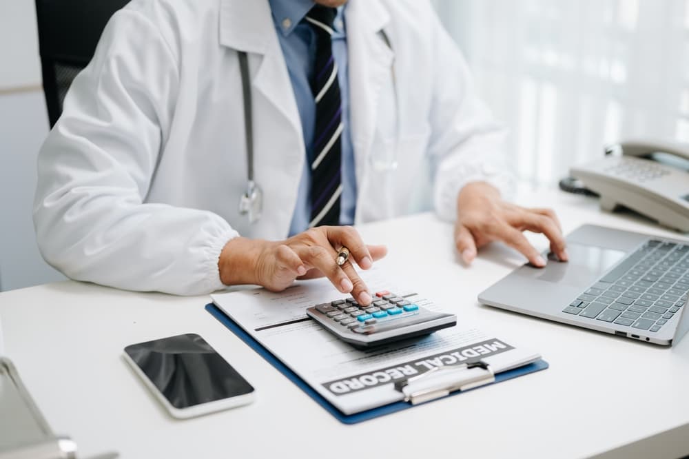 The concept of healthcare costs and fees: A doctor uses a calculator, smartphone, and tablet to assess medical expenses at a hospital.